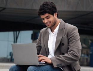 A man sitting on a bench, using a laptop, focused on his work in an outdoor setting