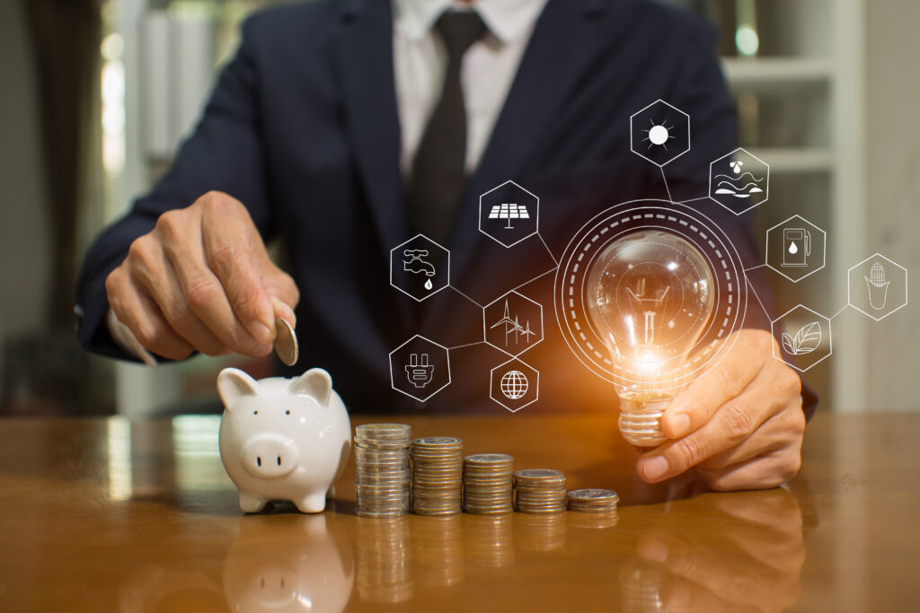 Person in a suit placing a coin into a piggy bank, with stacked coins and a glowing light bulb surrounded by business and technology icons in the foreground